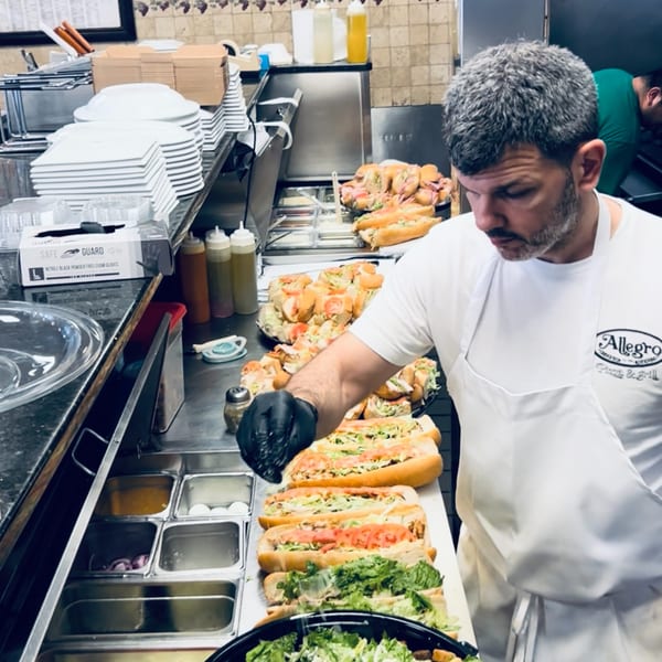Allegro Pizza chef in a white apron preparing sandwiches with fresh ingredients in a kitchen.