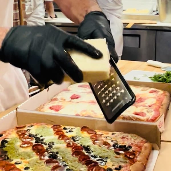 A chef in black gloves grates cheese over a freshly baked Allegro Pizza with various toppings.