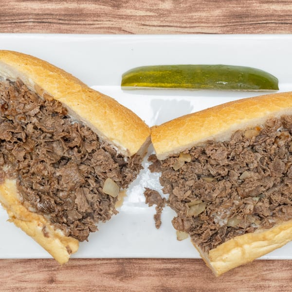 Allegro Pizza's cheesesteak sandwich, cut in half, with a pickle spear on a white plate.