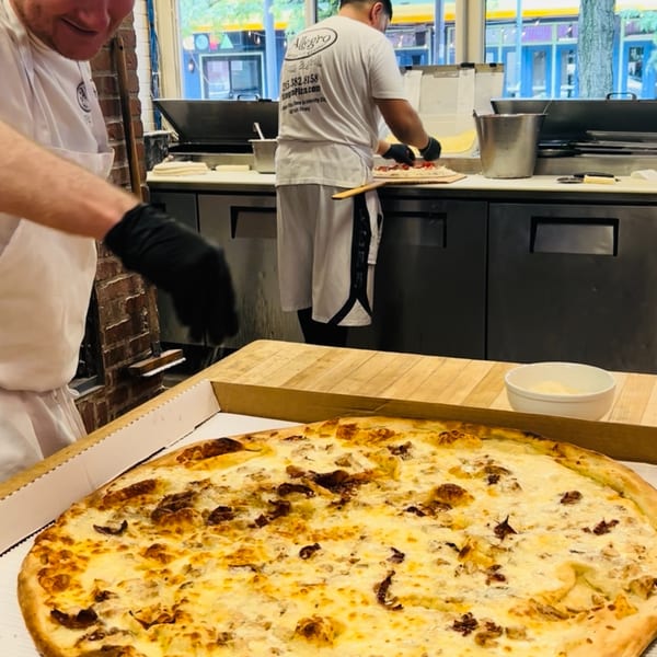 Allegro Pizza chefs preparing pizzas in a kitchen, with a large white pizza in the foreground.